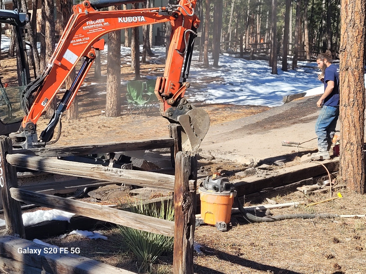 A mini excavator near a wooden fence, with a worker nearby, in a snowy forest area.