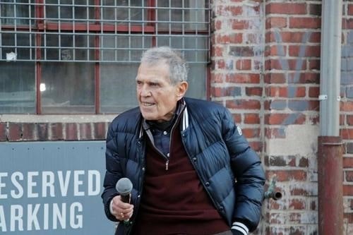 An older man stands near a brick wall, holding a microphone, wearing a jacket and speaking, with a sign that says "Reserved Parking" nearby.