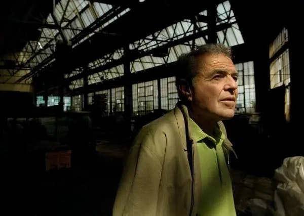 An older man stands in a dimly lit industrial space, gazing thoughtfully with light streaming through large windows.