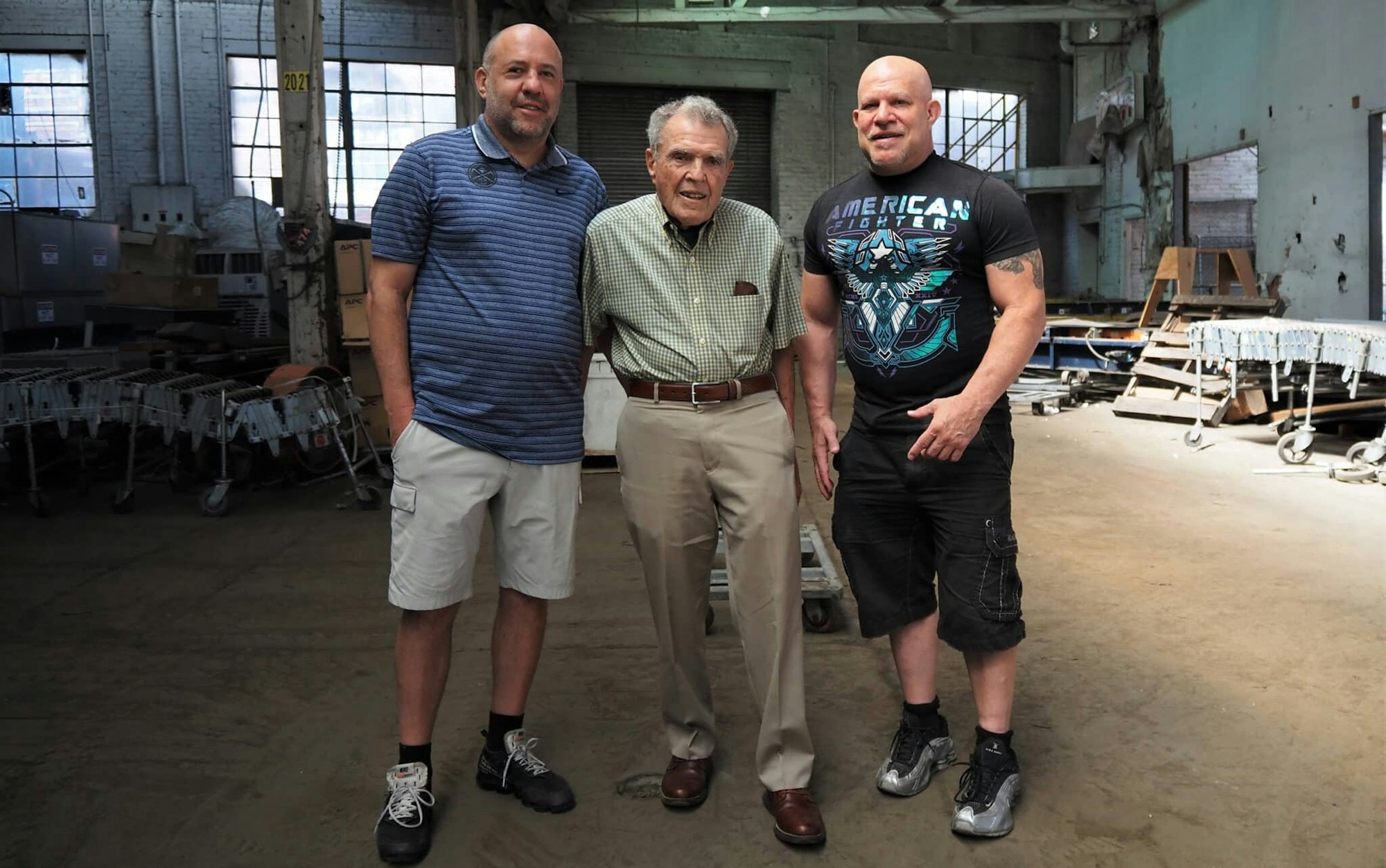 Three men stand together in an industrial setting, with one elderly man in the center, smiling for the photo.