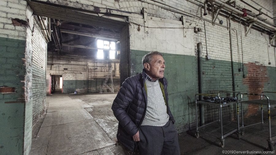 An older man stands in an empty, industrial-looking room with brick walls and large windows, suggesting a renovation or history.