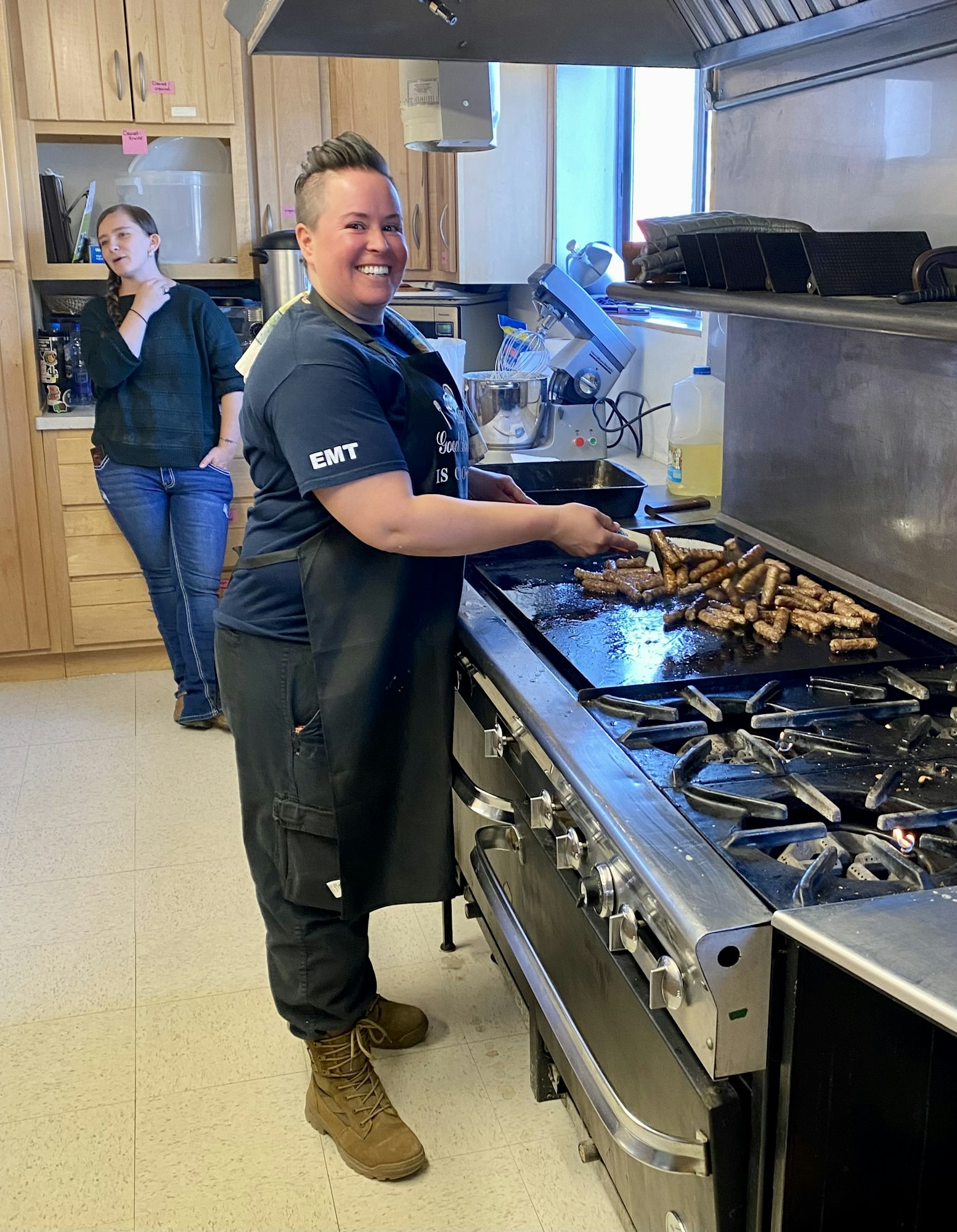 Person cooking sausages on a griddle in a kitchen, smiling, with another person in the background.