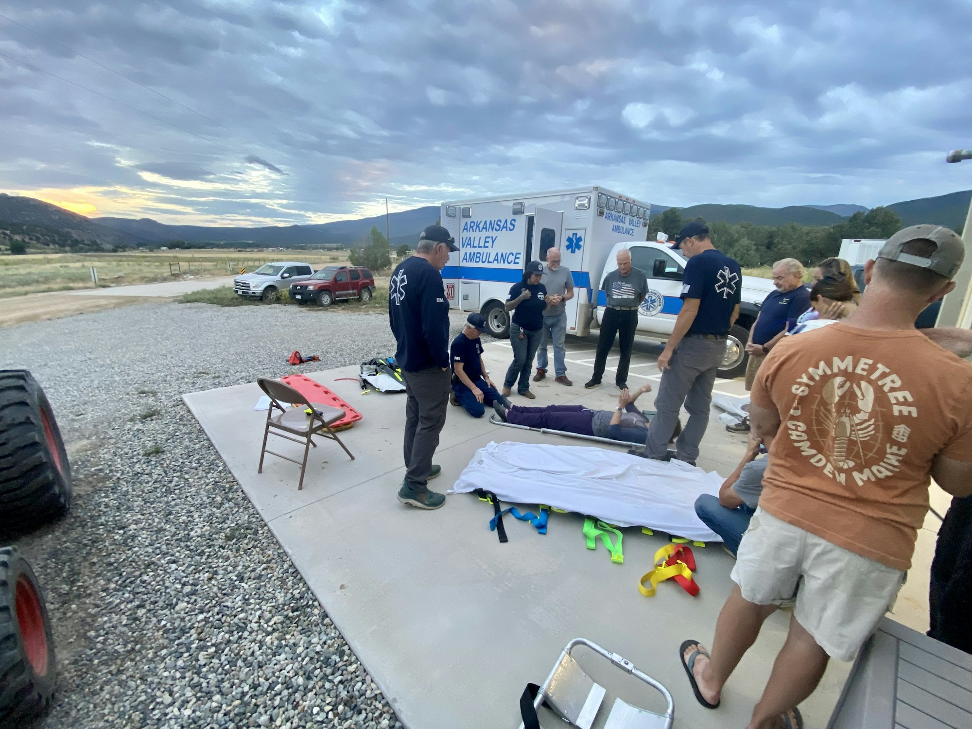 A group of people are gathered around a person on the ground near an ambulance, seemingly for a medical exercise or training.