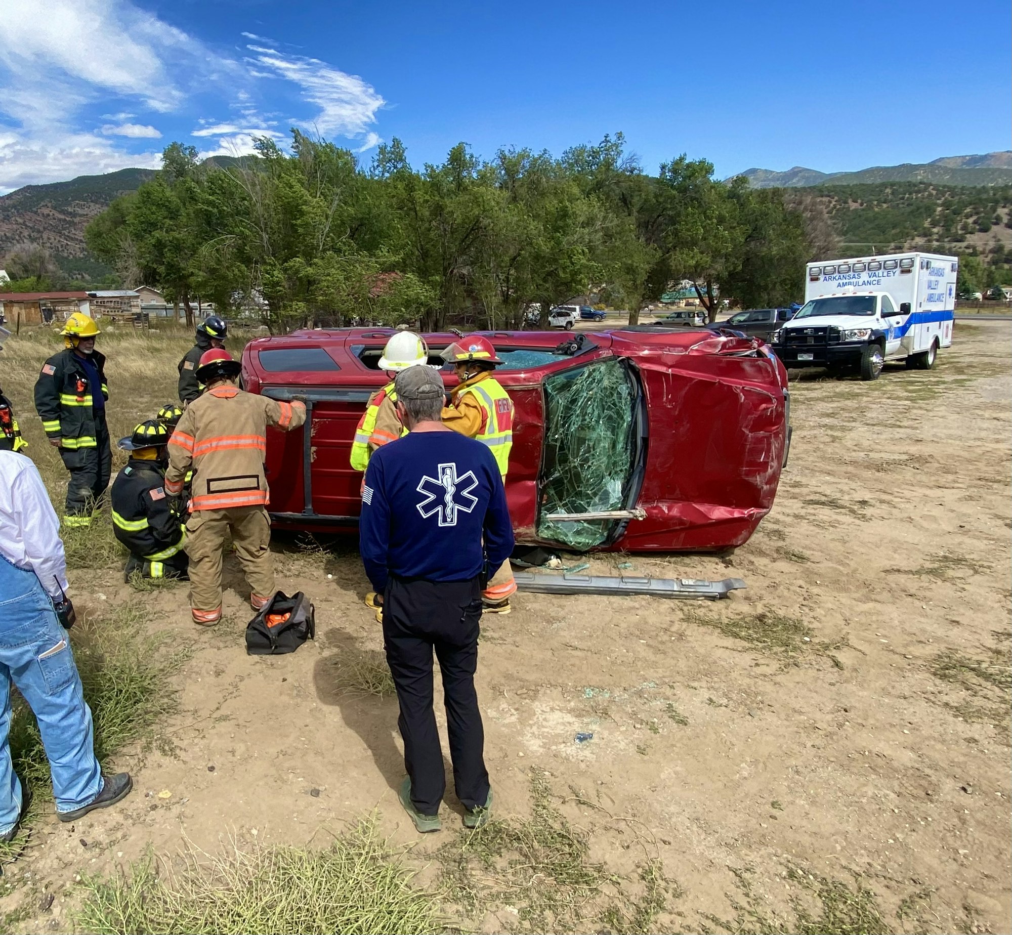 Emergency responders and firefighters assess a red overturned vehicle. An ambulance is on the scene.