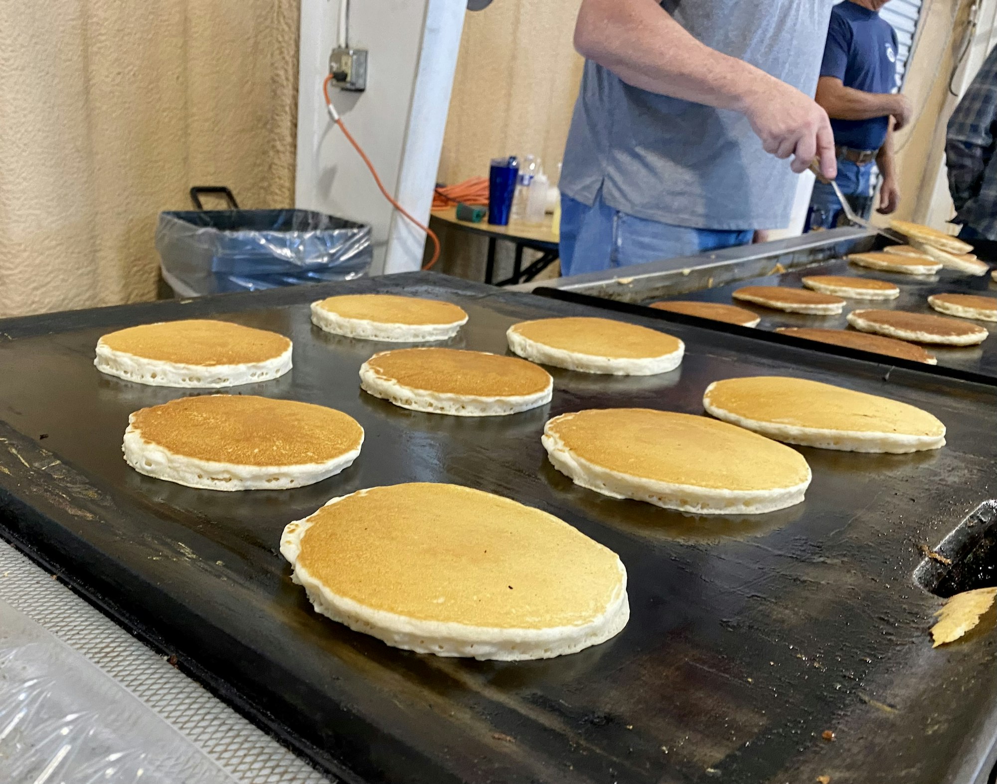 Pancakes cooking on a large griddle, with a person flipping them.