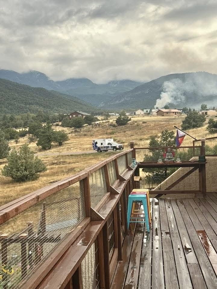 A wooden deck with stools, overlooking a field and mountains; smoke rises from a distant building and an ambulance is nearby.