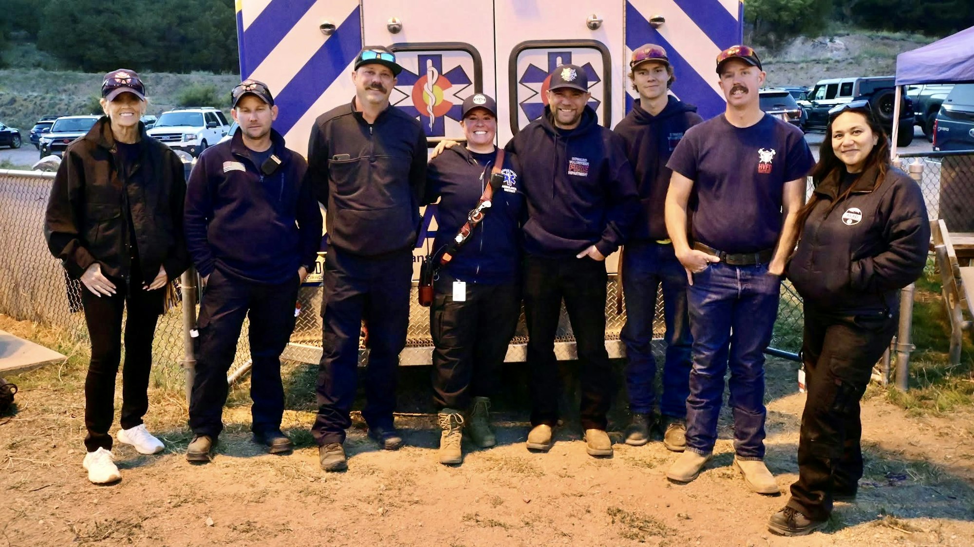 A group of eight people in casual outdoor wear standing in front of a vehicle with emergency symbols.