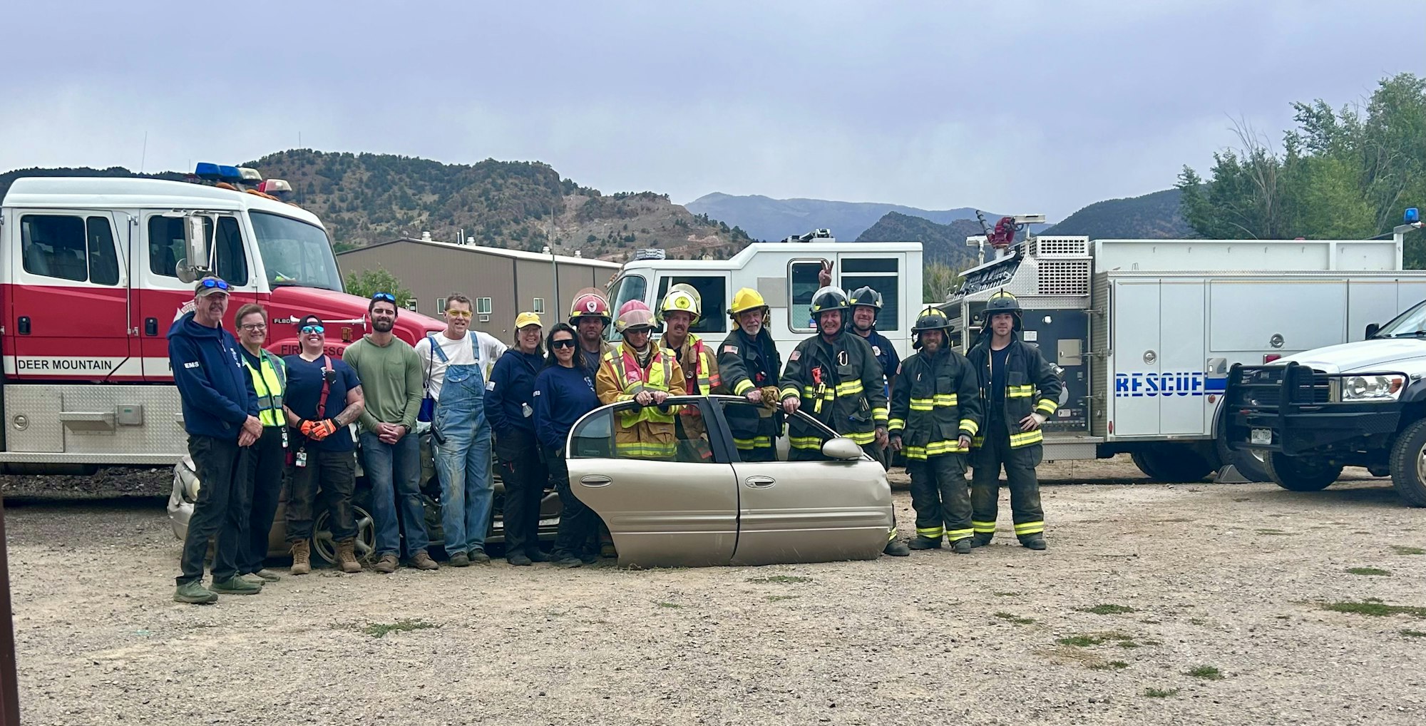A group of firefighters and emergency personnel posing in front of fire and rescue trucks, holding a car door.