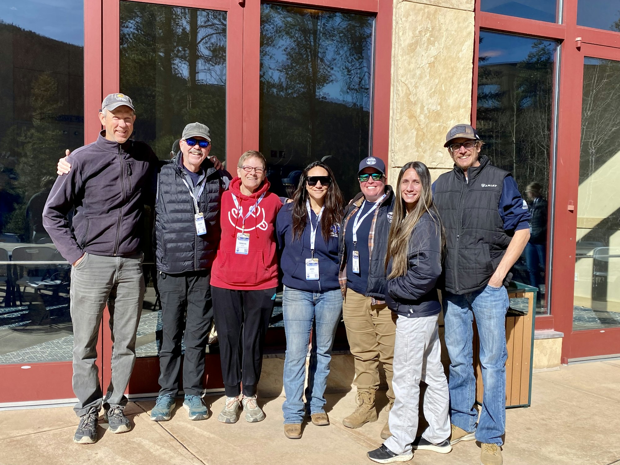 A group of seven people, wearing casual outdoor clothing, standing in sunlight in front of a building with large windows.