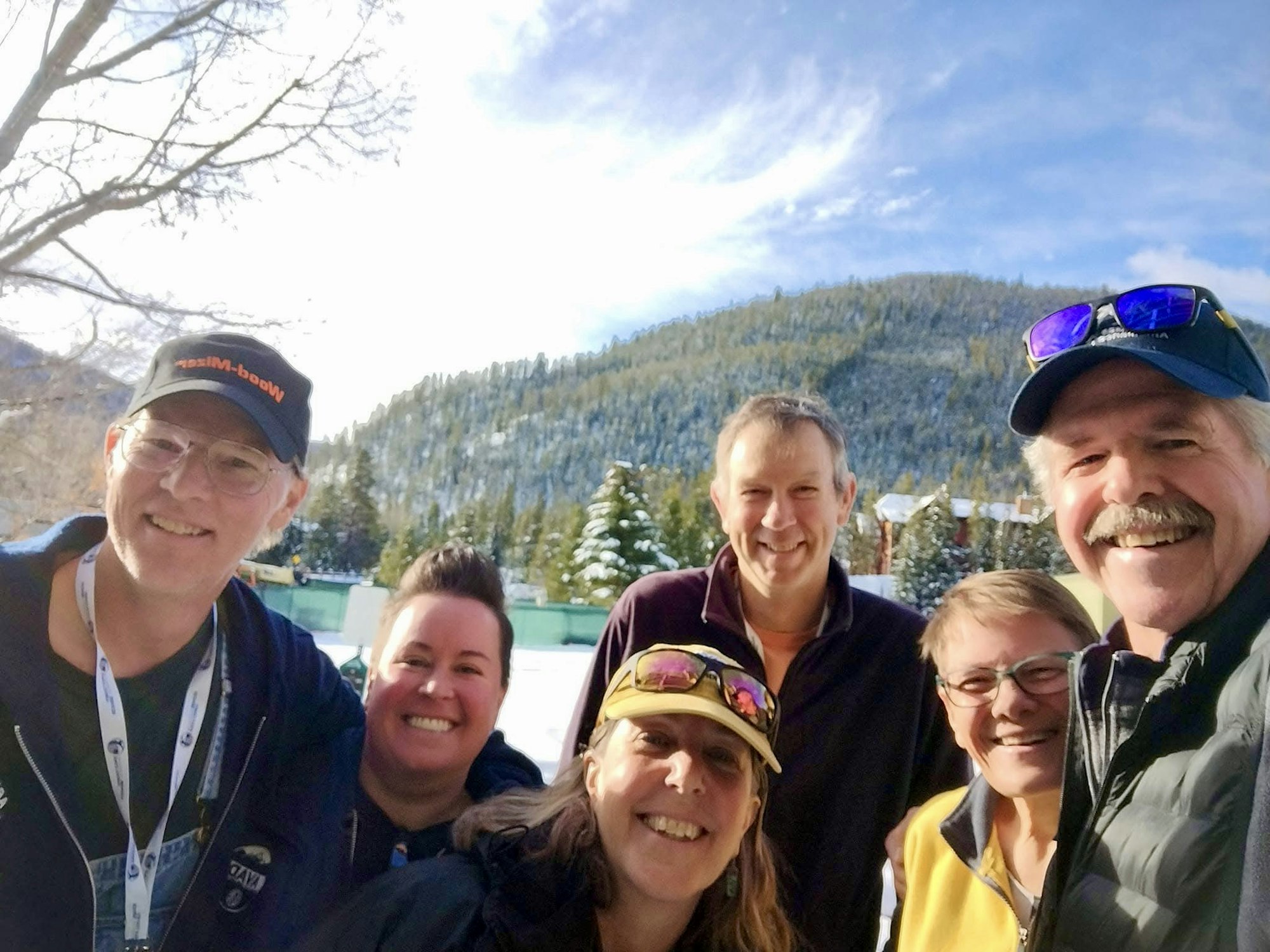 A group of six smiling people outdoors, snowy mountains in the background.