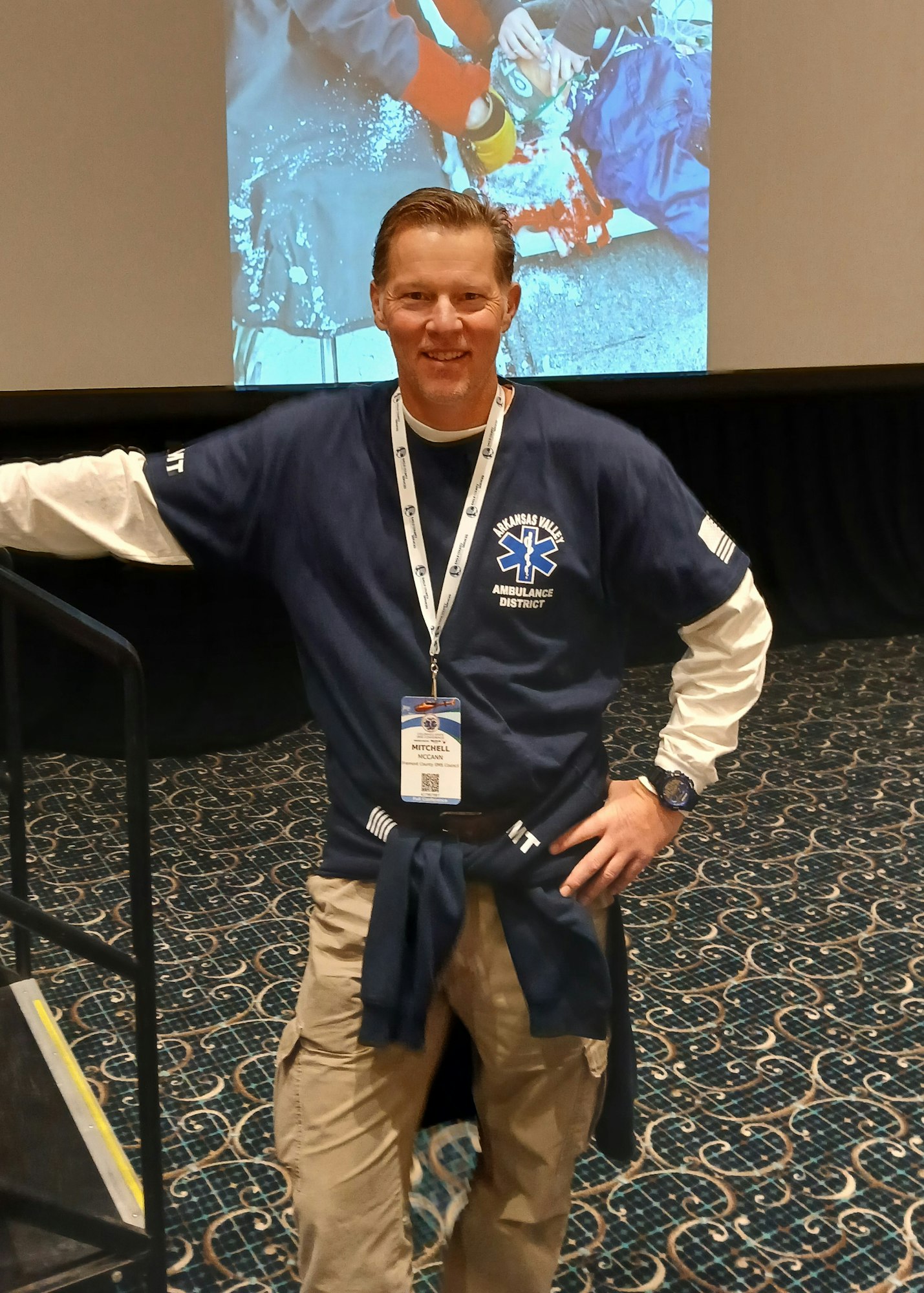 A person in an "Arkansas Valley Ambulance" shirt stands smiling indoors, wearing a lanyard and badge.