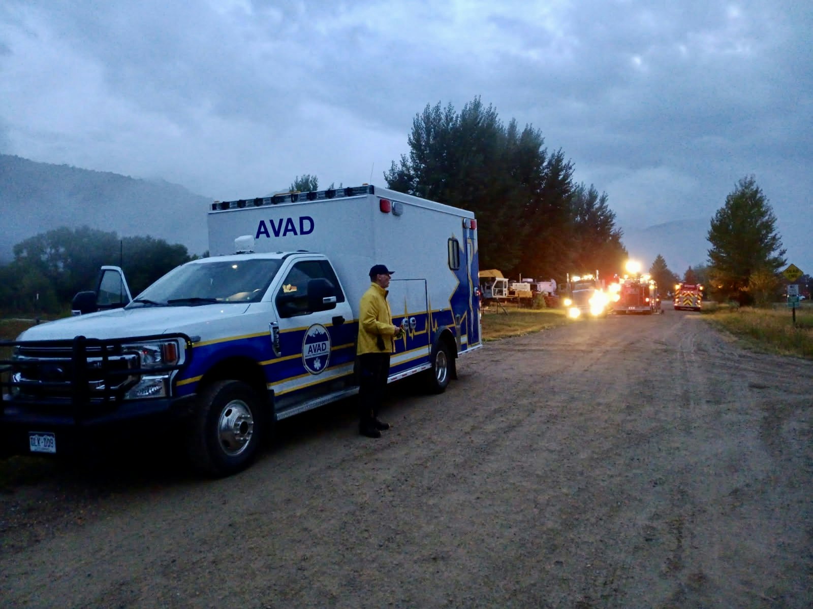 Emergency vehicles and personnel on a dirt road, with trees and a cloudy sky in the background.