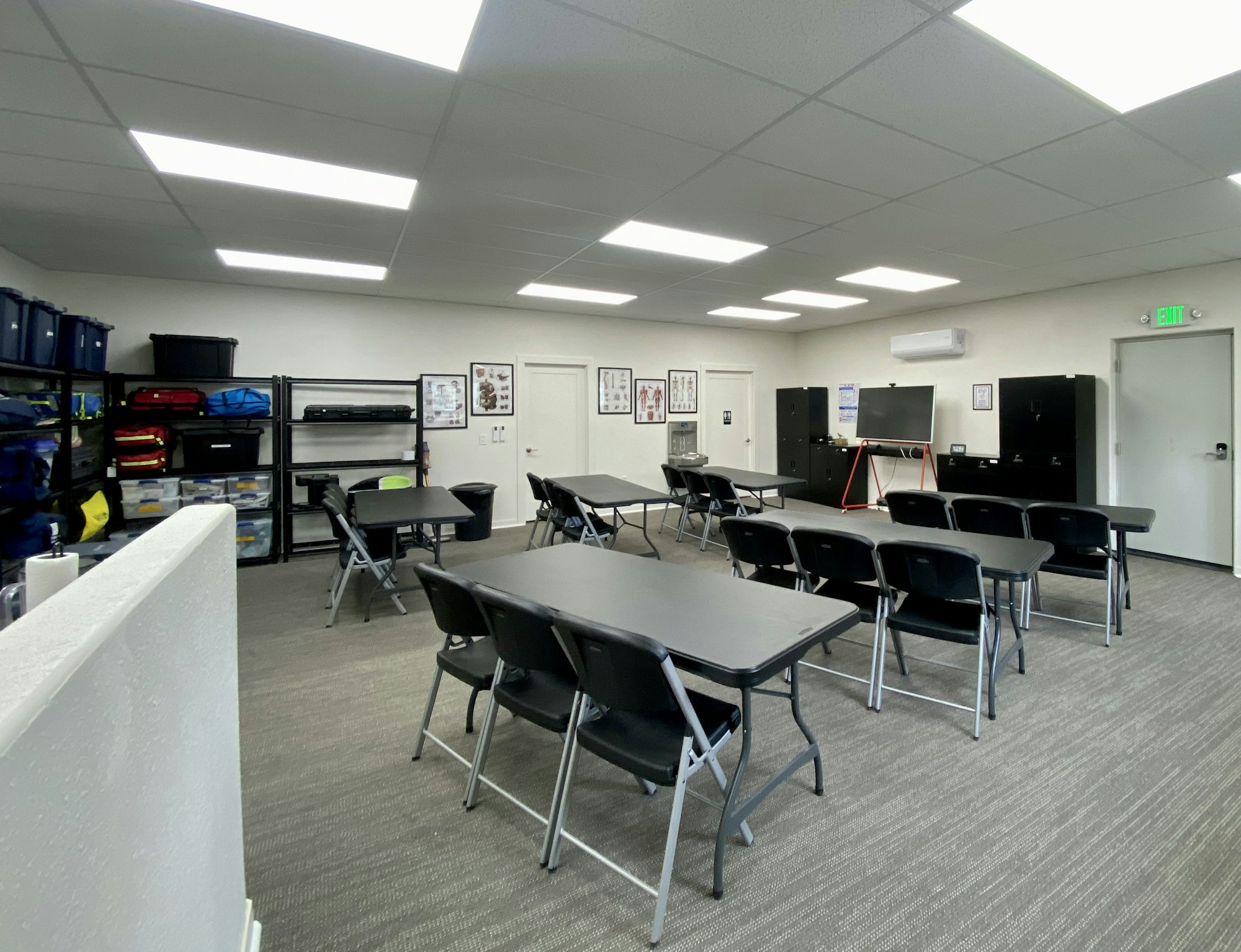 A well-lit classroom with folding tables, chairs, storage shelves, educational posters, and a black screen on a stand.