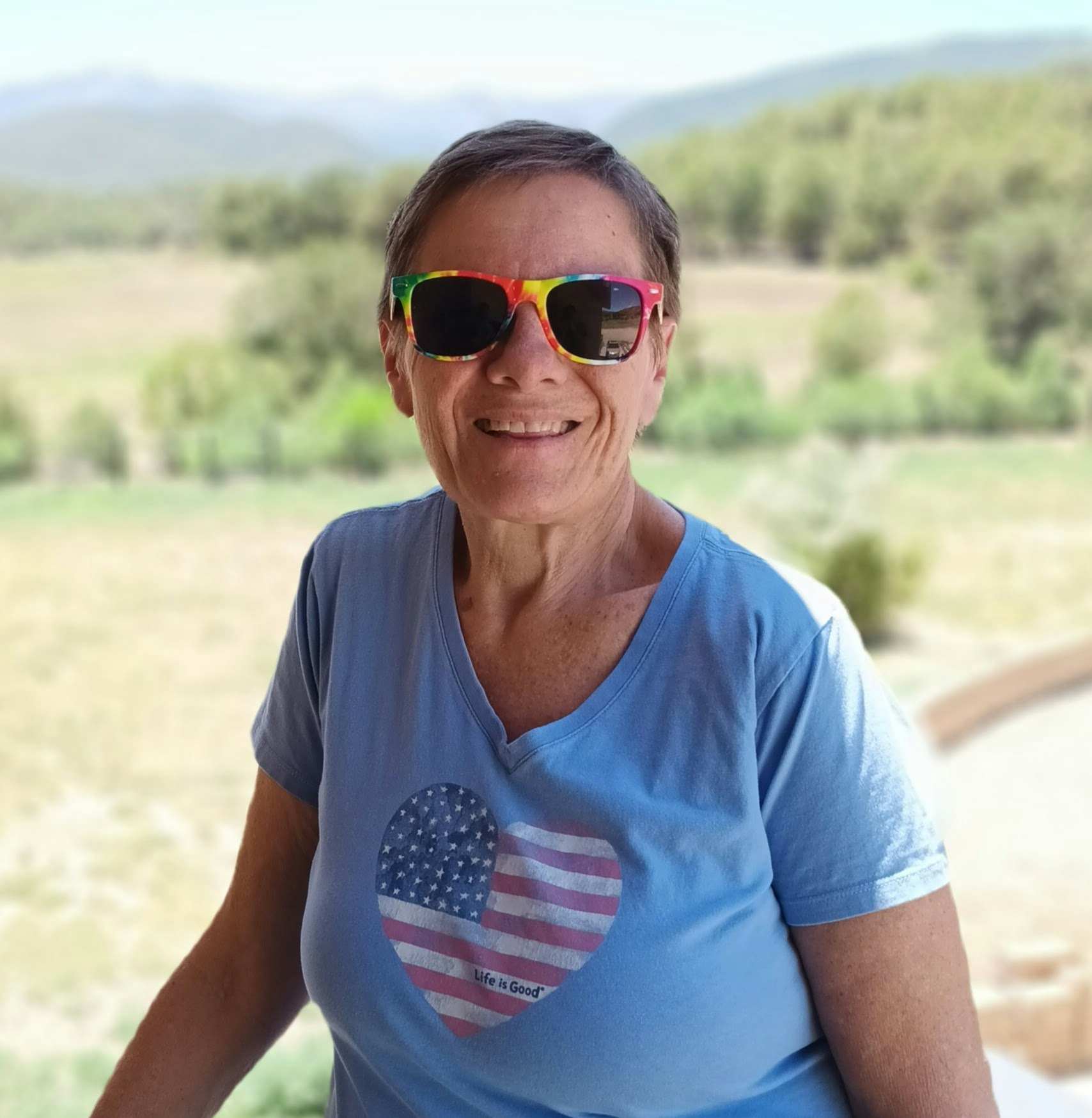 A person wearing rainbow sunglasses and a blue shirt with a heart design stands outdoors in a scenic setting.