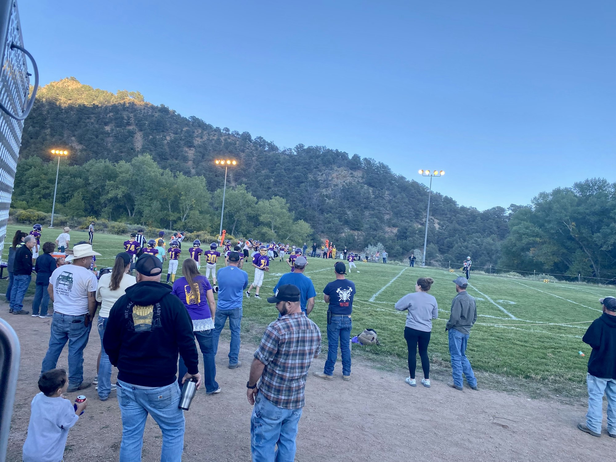 A group of people watching a football game on a grassy field with mountains and trees in the background.
