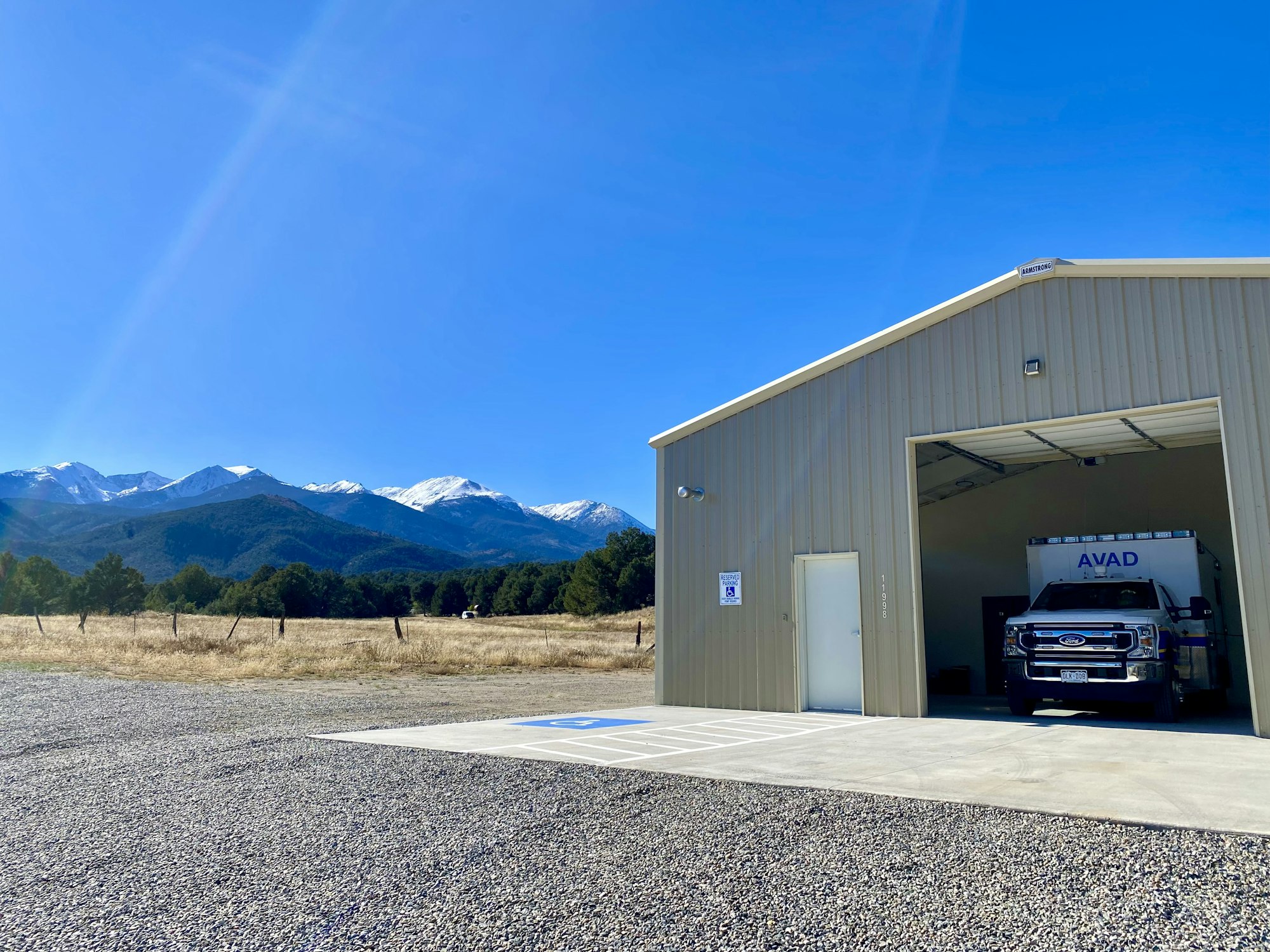 A large garage with a truck inside, gravel foreground, mountains and trees in the background under a clear blue sky.
