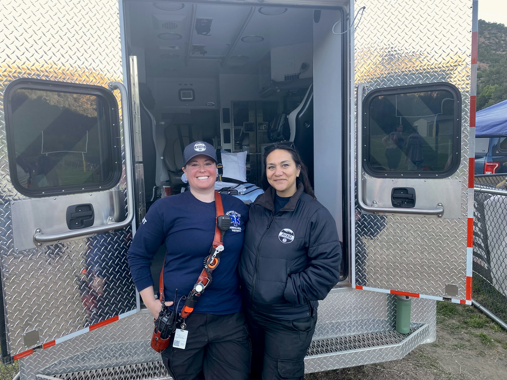 Two people in uniform smiling in front of an open emergency vehicle.