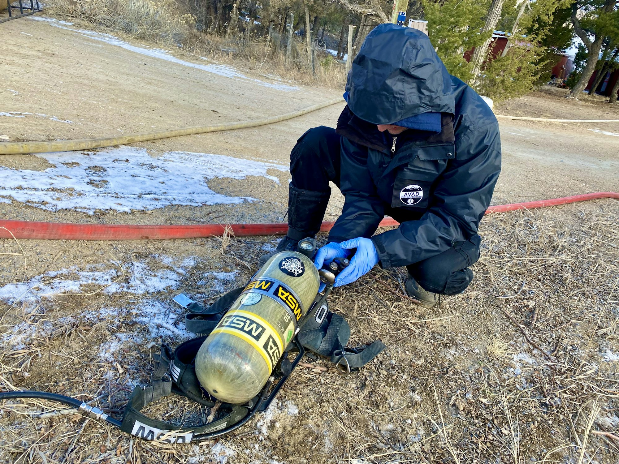 A person in outdoor gear adjusts an MSA air tank, wearing gloves, on a snowy, grassy area with hoses nearby.