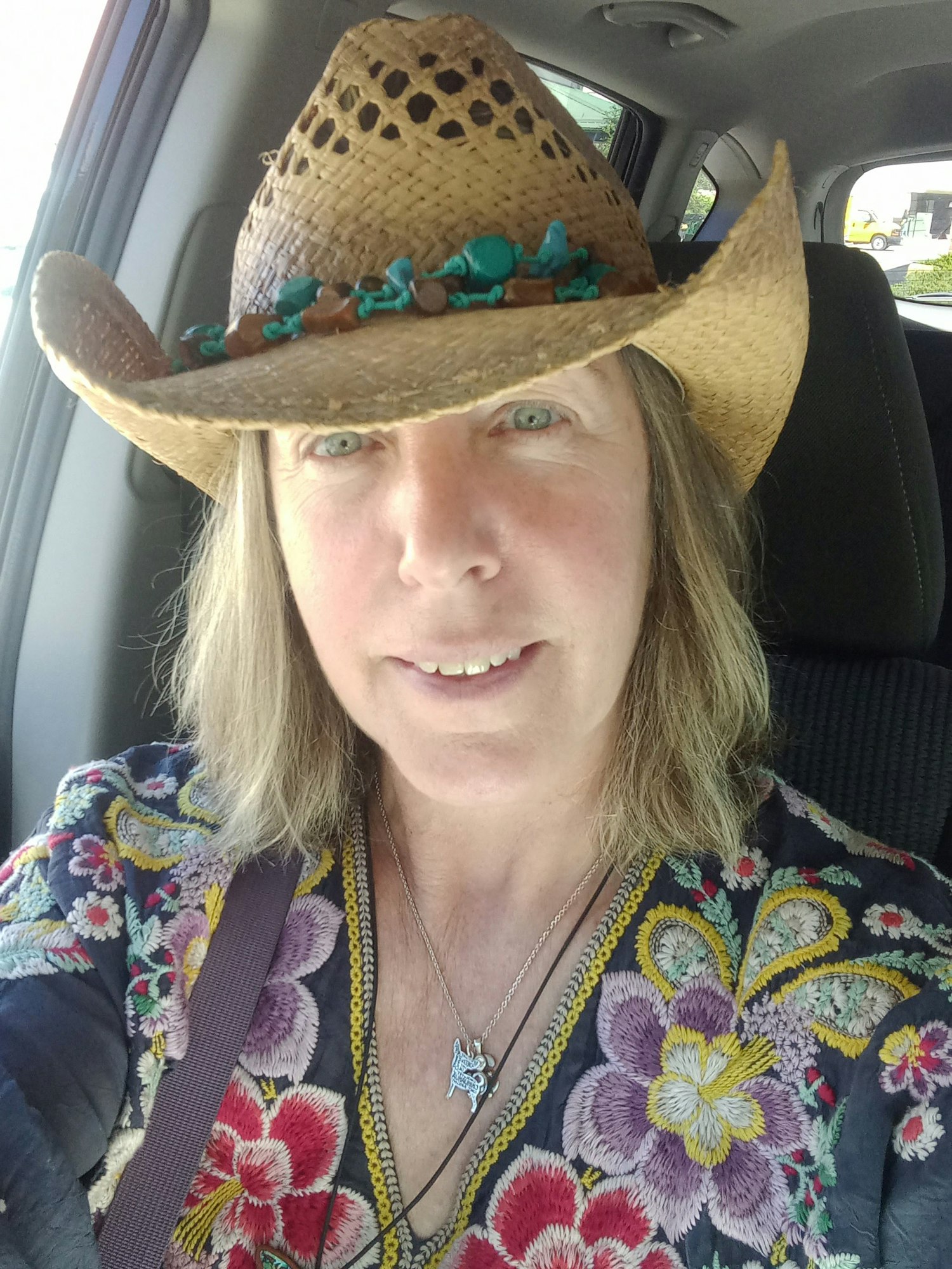 A person in a colorful floral top and straw hat with turquoise embellishments, sitting inside a vehicle.