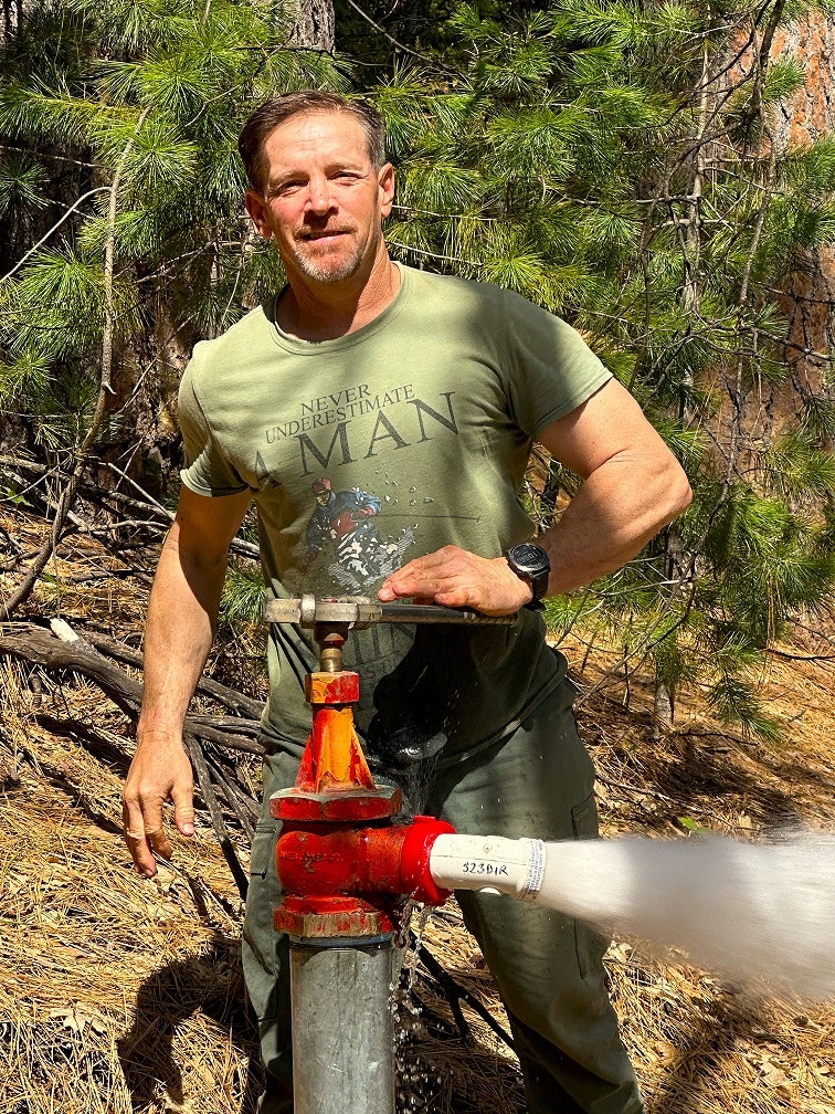 A man operates a fire hydrant, water gushing out, in a forest setting.