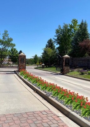 A gated community entrance with brick pillars, black metal gates, and a landscaped median with red flowers, surrounded by trees.