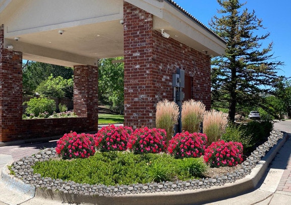 Brick structure with a flower bed featuring red blooms, green shrubs, and ornamental grasses, surrounded by a stone border.