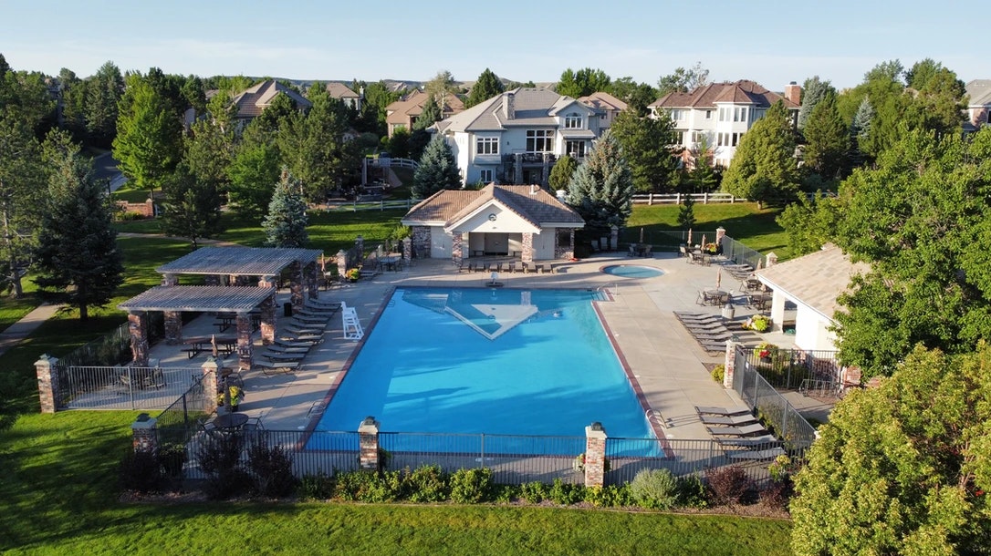 A clear swimming pool with lounge chairs, surrounded by greenery and residential houses in the background.