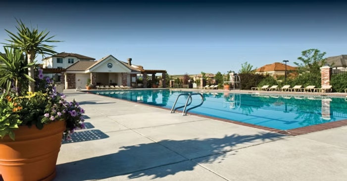 Outdoor swimming pool with a diving board, surrounded by lounge chairs and plants, in front of a clubhouse.