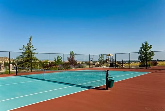 Tennis court with a net, surrounded by a fence. Playground equipment and trees in the background. Blue sky above.