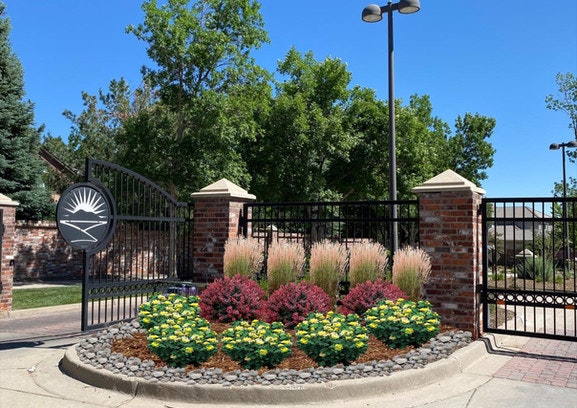 A gated entrance with brick pillars, decorative gardens, and a metal gate featuring a sunburst design, surrounded by trees.