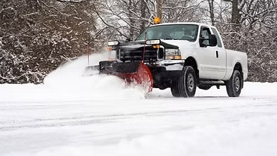 A white pickup truck with a snowplow attached, clearing snow from a road in a snowy landscape.