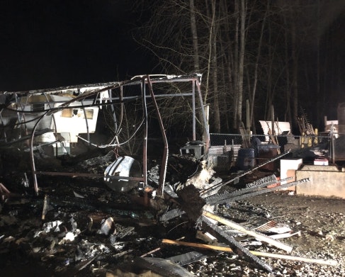 The image shows a damaged structure, likely from a fire or collapse, surrounded by debris and trees in a dark setting.