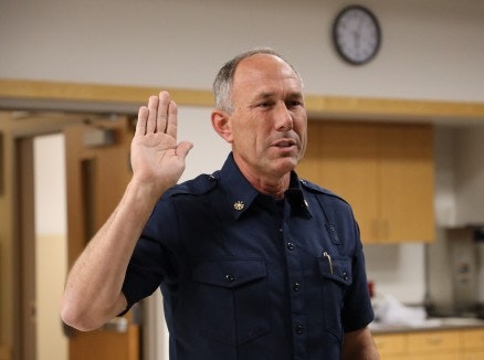 A person in a uniform is raising their right hand, possibly taking an oath, in a room with wooden furnishings and a clock.