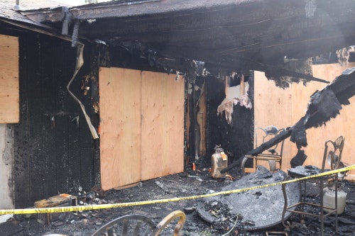 A charred building exterior with boarded windows, damaged structure, and debris, indicating a recent fire incident.