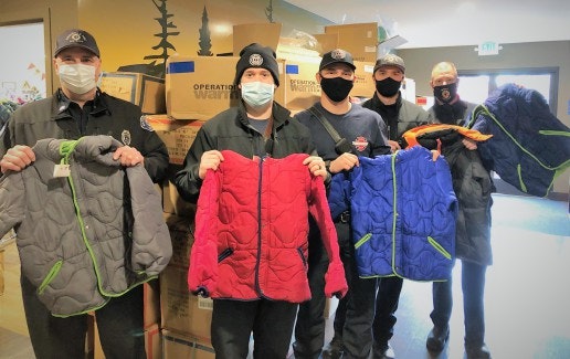 A group of masked individuals holding colorful jackets in front of boxes, likely part of a community outreach or donation effort.