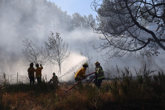 Firefighters are battling a wildfire, using equipment to manage smoke and flames amidst a wooded area.