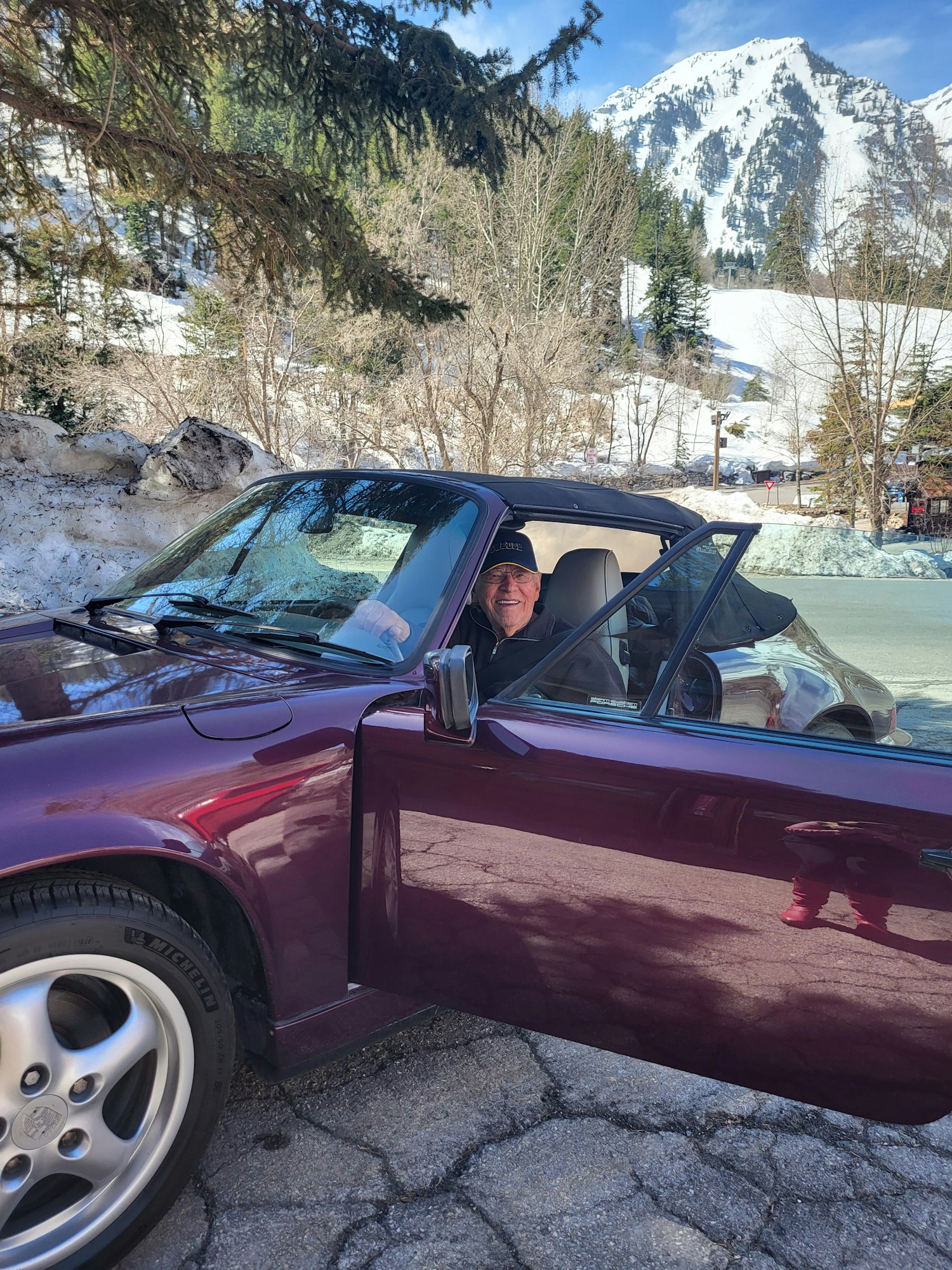 Maroon car with open door, person inside, snowy mountain background. (Sideways image)