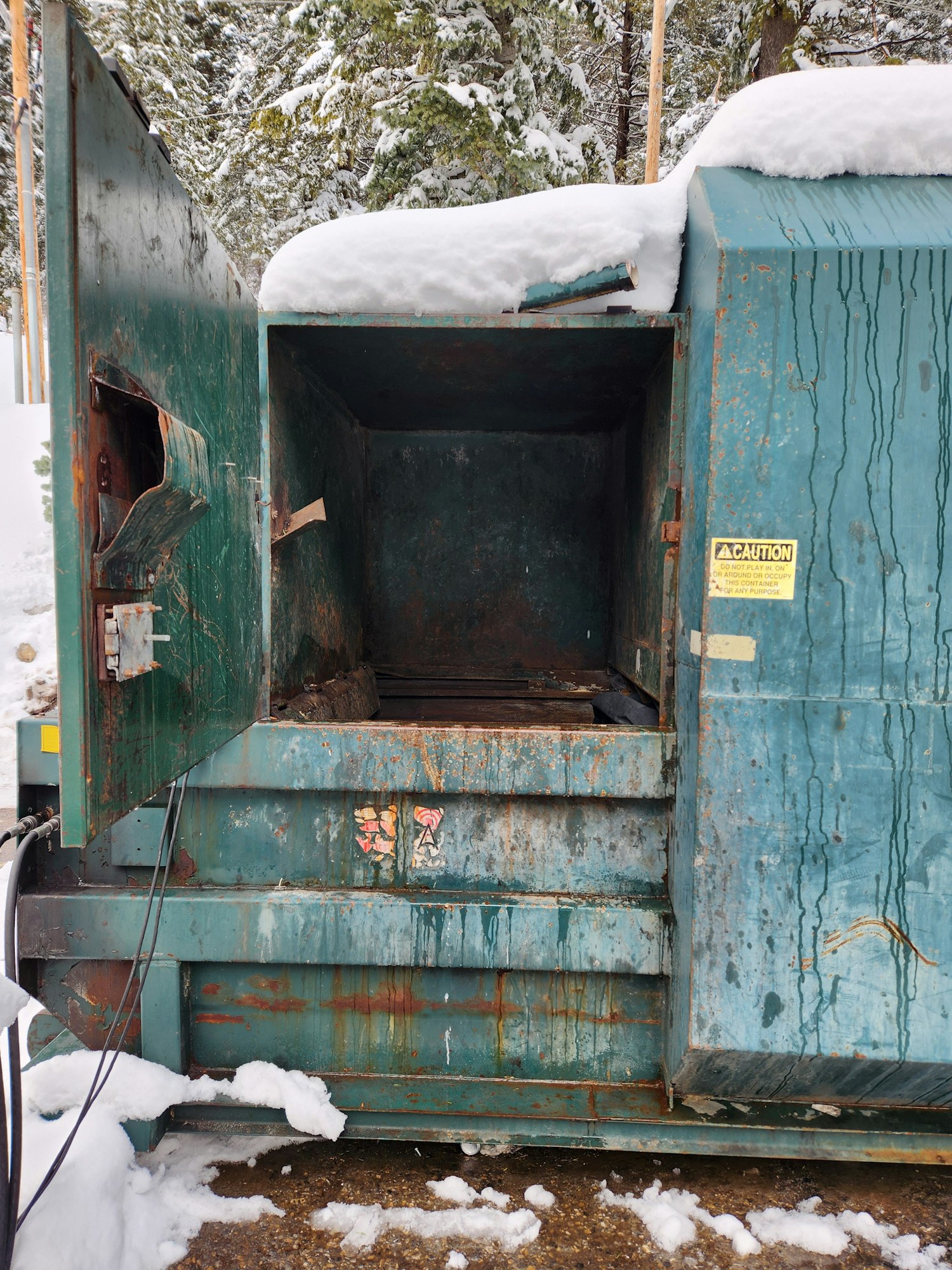 An empty, rusty dumpster with its lid open, surrounded by snow, turned sideways in the image.