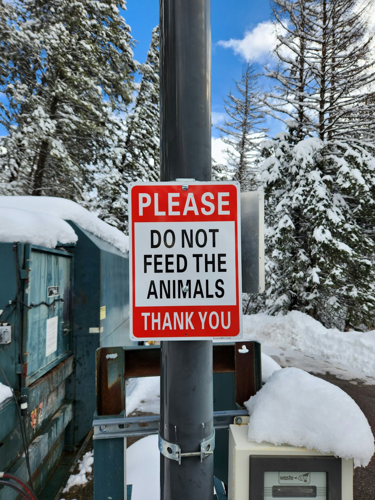 A sign reading "PLEASE DO NOT FEED THE ANIMALS THANK YOU" in a snowy environment with trees and an electrical box.