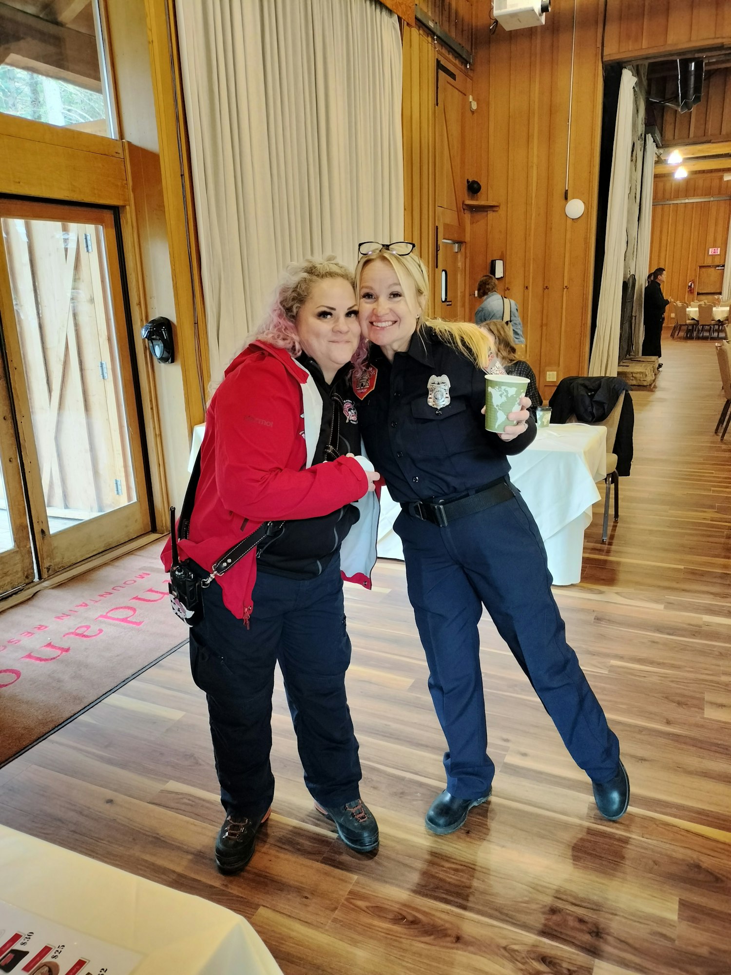 Two women smiling and embracing, one in a red jacket and the other in a police uniform, in a room with wooden interiors.