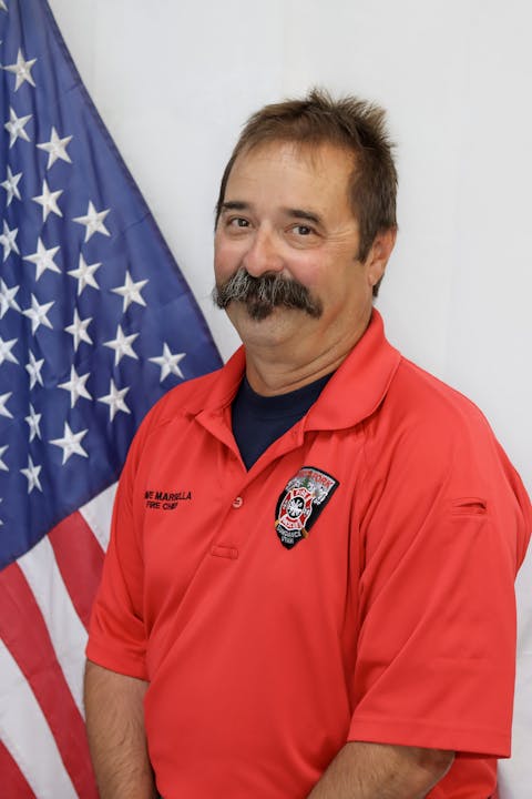 A man in a red firefighter shirt smiling in front of an American flag.