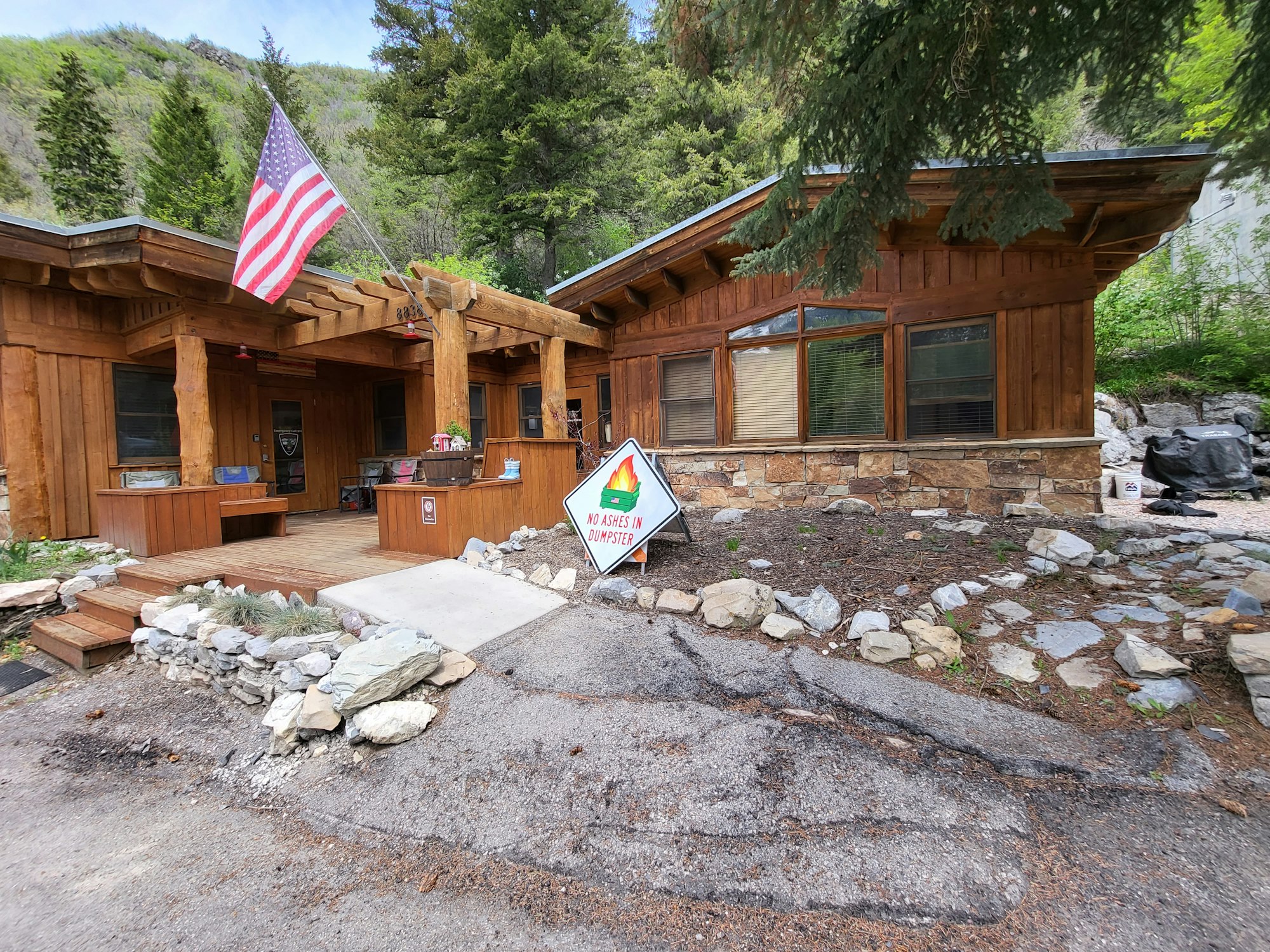 A rustic wooden building with an American flag, surrounded by nature. A sign reads "No ashes in dumpster."