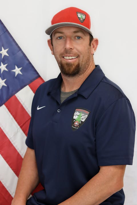 A man in a polo and cap stands before the American flag.