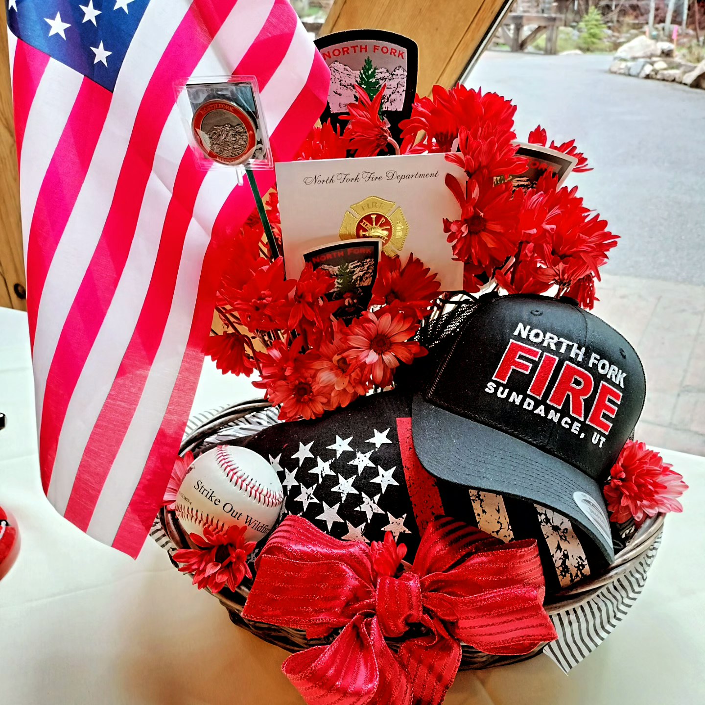 A patriotic display with an American flag, artificial red flowers, a firefighter cap, a baseball, and sundry items in a basket.