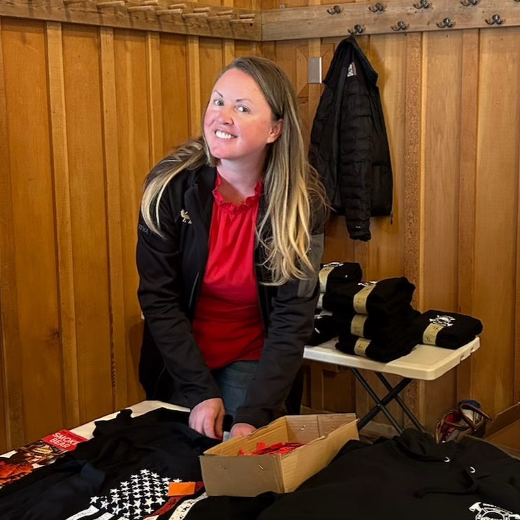 A smiling woman with a table with merchandise—t-shirts, caps, and brochures in a wooden-walled room.