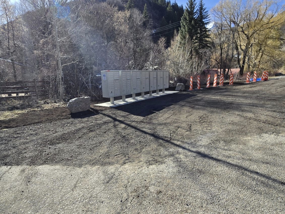 Outdoor mailbox cluster beside a dirt road, surrounded by trees and caution barriers.