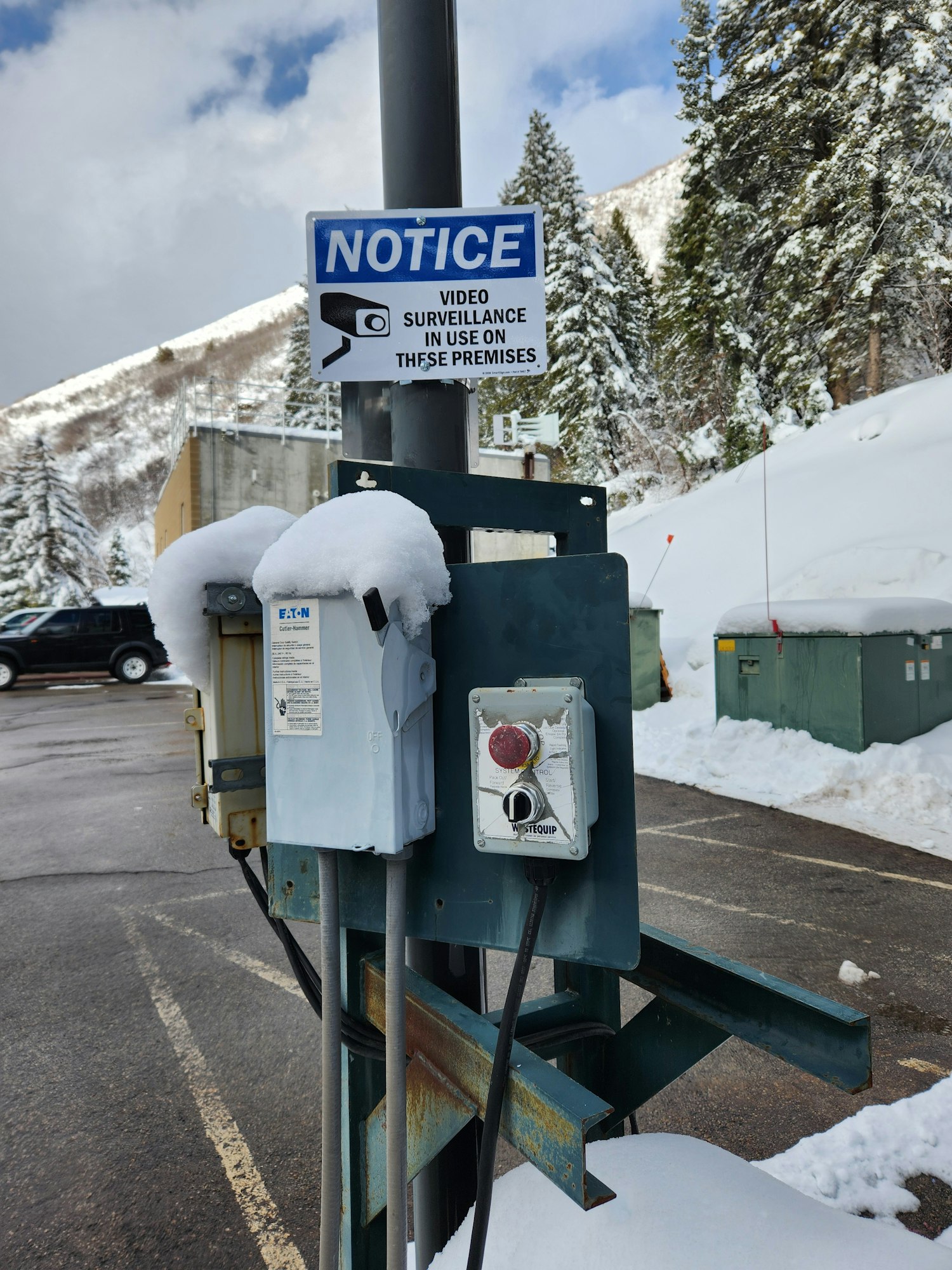 Snow-covered utility box with surveillance sign, snowy trees, and mountain backdrop. (Image is rotated.)