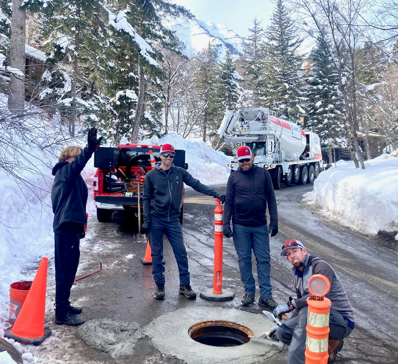 Four workers near an open manhole with a truck and snow-covered trees in the background.