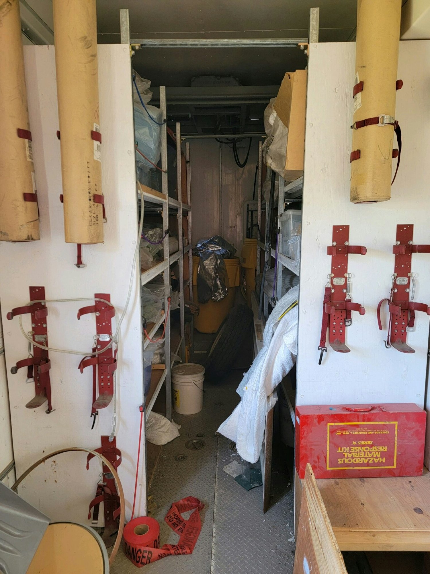 A cluttered storage room with shelves, safety gear, and a "Hazardous Material Response Kit" box.