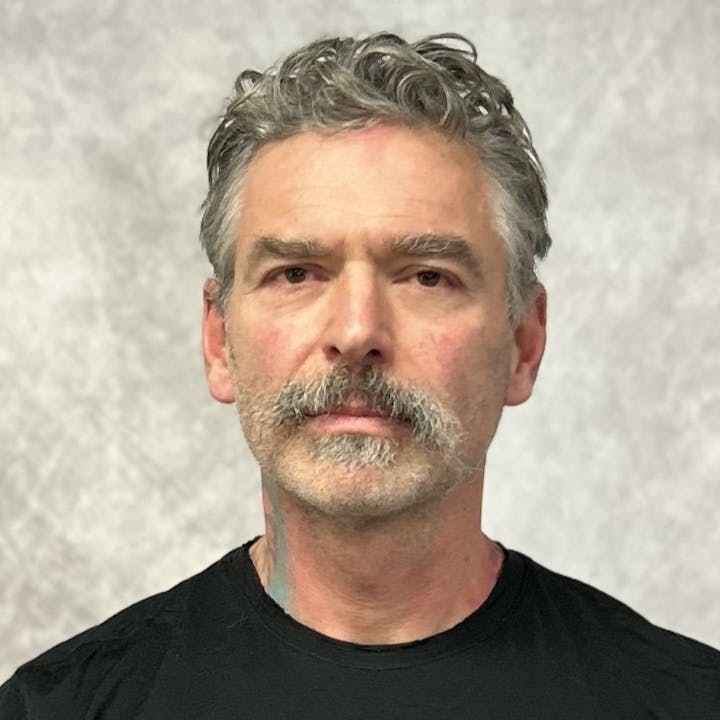A person with a mustache and gray hair wearing a black shirt against a neutral background.